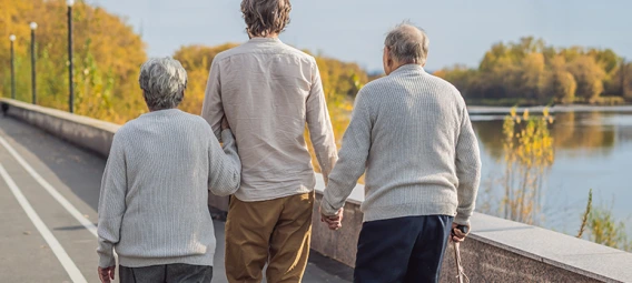 Adult caregiver walking with two elderly individuals along waterfront path in autumn, holding hands for support and companionship