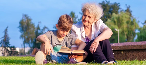 An older woman and young boy sitting on grass outdoors, reading a book together.
