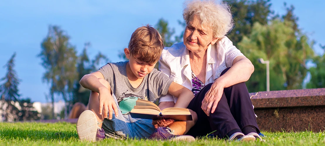 An older woman and young boy sitting on grass outdoors, reading a book together.