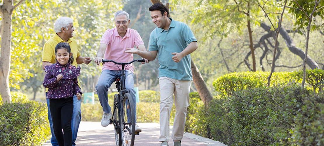 Three generations walk together outdoors — a young girl, an older man on a bike, and a younger man, on a tree-lined path.