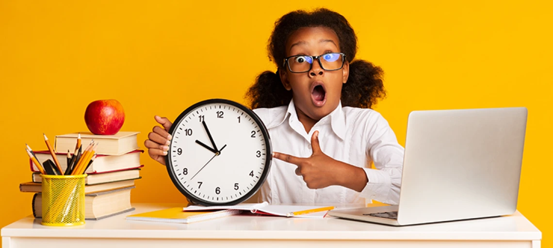 : A young girl with glasses points at a large clock while sitting at a desk with a laptop.