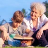 An older woman and young boy sitting on grass outdoors, reading a book together.