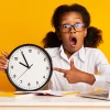 : A young girl with glasses points at a large clock while sitting at a desk with a laptop.